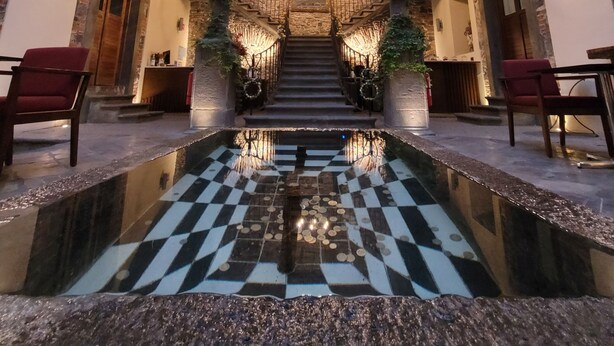A grand staircase with a checkered floor leading to a balcony, surrounded by stone walls and furnished seating. - Milo Collection Hotel (Puebla)