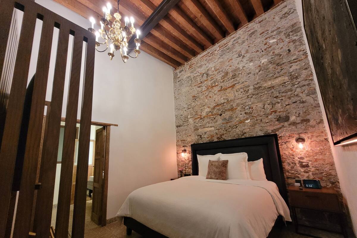 A bedroom with a stone wall, a wooden ceiling, a chandelier, a bed with white linens, and a nightstand with a clock.