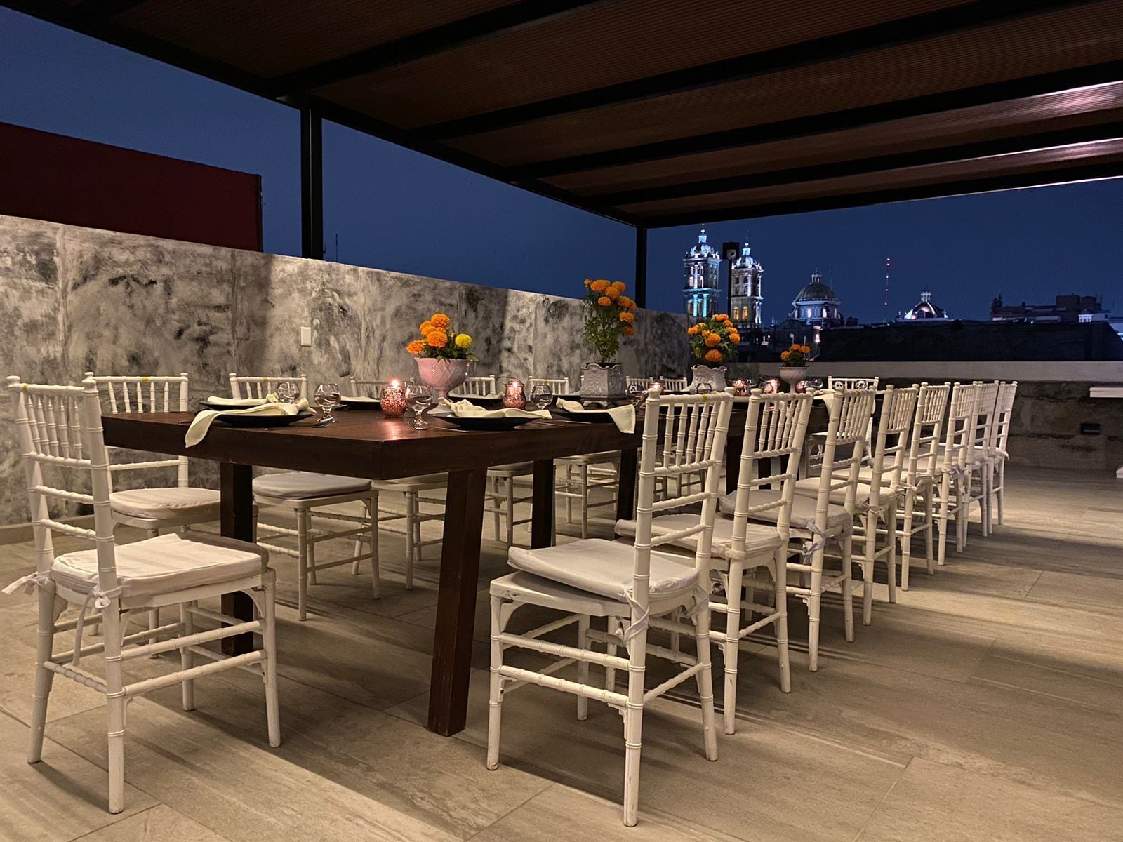 Uma sala de jantar com mesa posta para 12 pessoas, rodeada por cadeiras brancas, sob um alpendre coberto com vista noturna para a cidade.