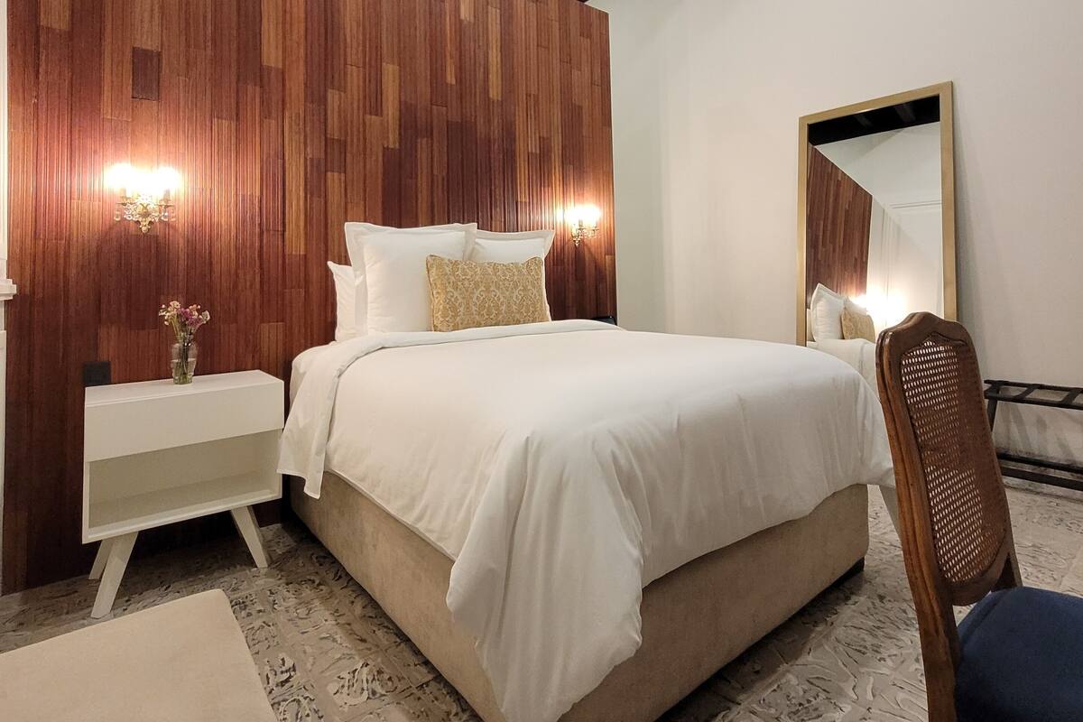 A bedroom with a wooden panel wall, a bed with white bedding, a nightstand with a vase, and a chair.