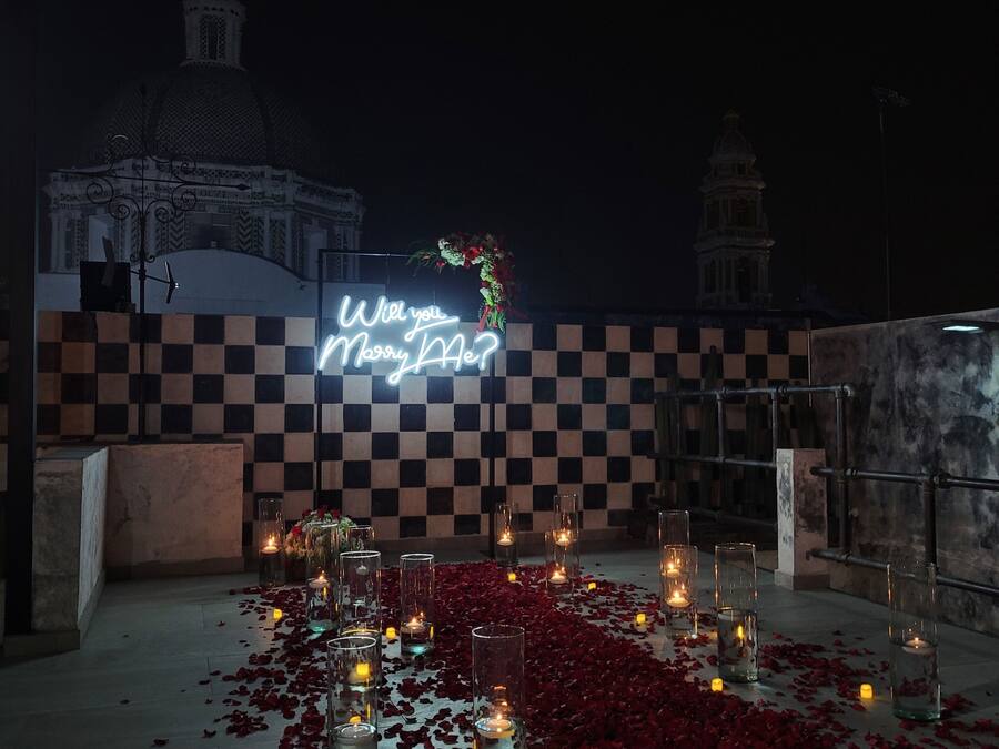 A romantic outdoor setting with a neon sign reading "Will you marry me?" surrounded by candles and rose petals.