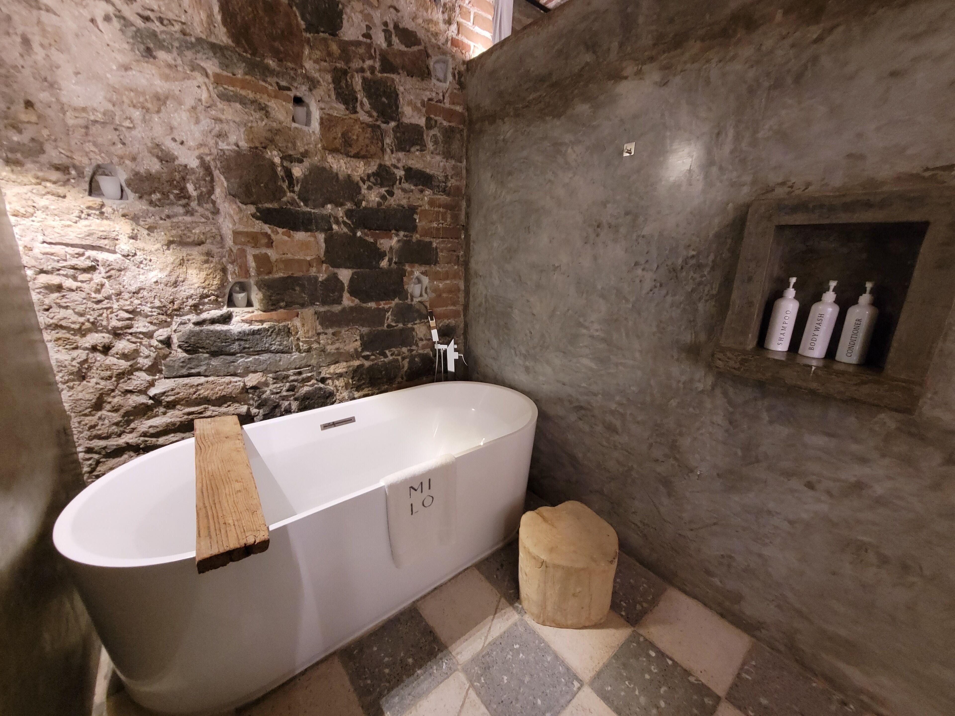 Une salle de bain aux murs de pierre, avec une baignoire sur pieds, un tabouret en bois et une étagère contenant des articles de toilette.