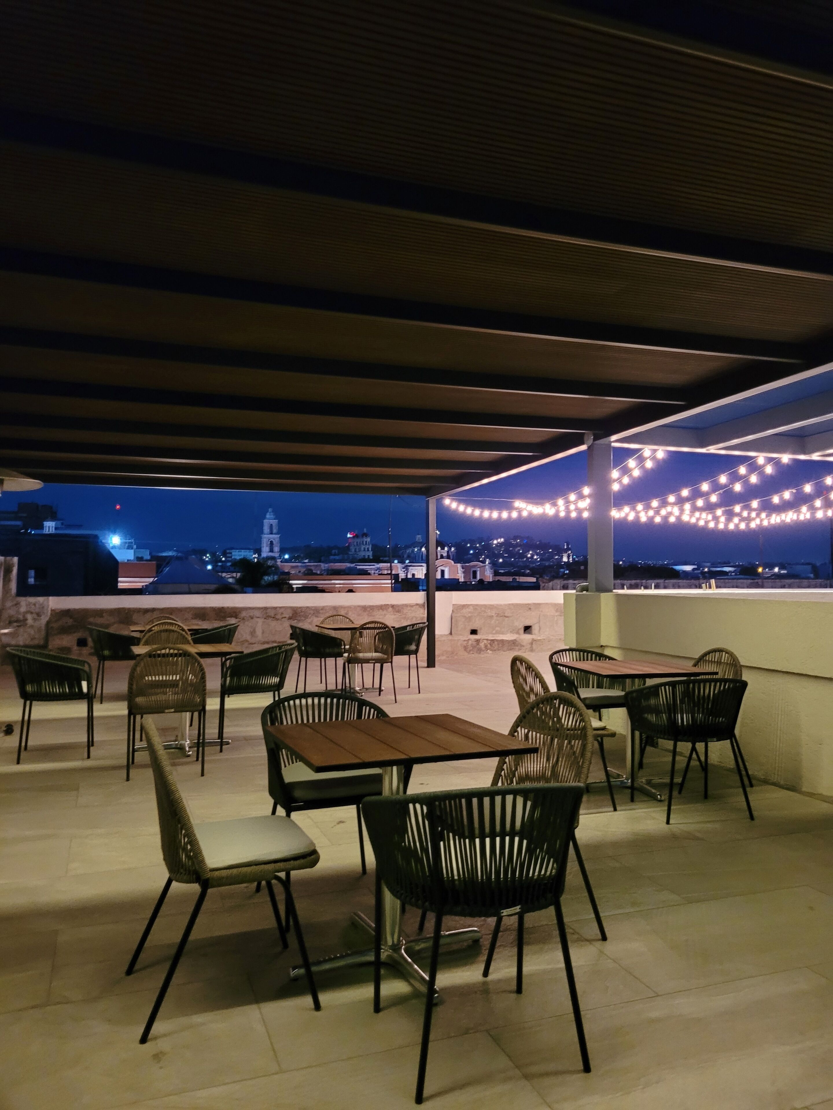 An outdoor patio with tables and chairs, string lights, and a view of the cityscape at dusk.