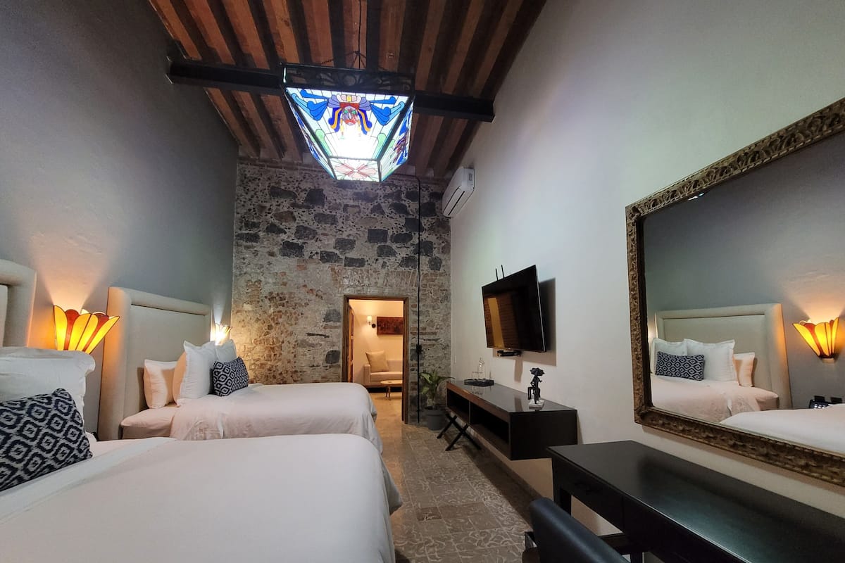 A hotel room with two beds, a TV, a mirror, and a stone wall.