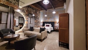 A hotel room with a stone wall, a bed with white linens, a wooden wardrobe, and a seating area with a sofa and chairs. - Milo Collection Hotel (Puebla)