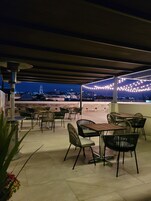 Un patio al aire libre con mesas y sillas, luces colgantes y vistas a un paisaje urbano al anochecer.