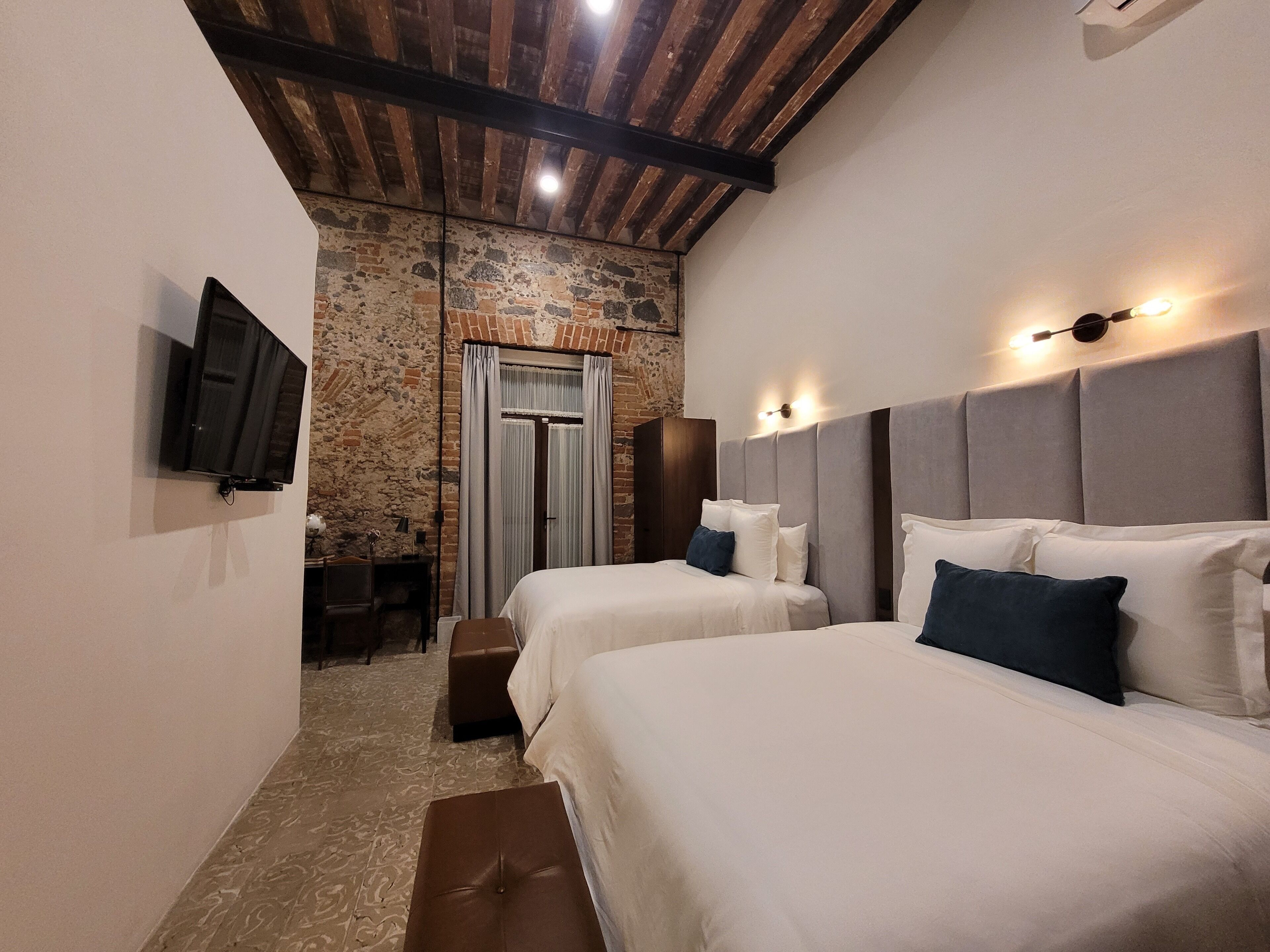 A bedroom with two beds, a stone wall, a wooden ceiling, and a TV mounted on the wall.