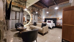 A hotel room with a stone wall, a bed with white bedding, a sofa, a coffee table, and a TV mounted on the wall. - Milo Collection Hotel (Puebla)