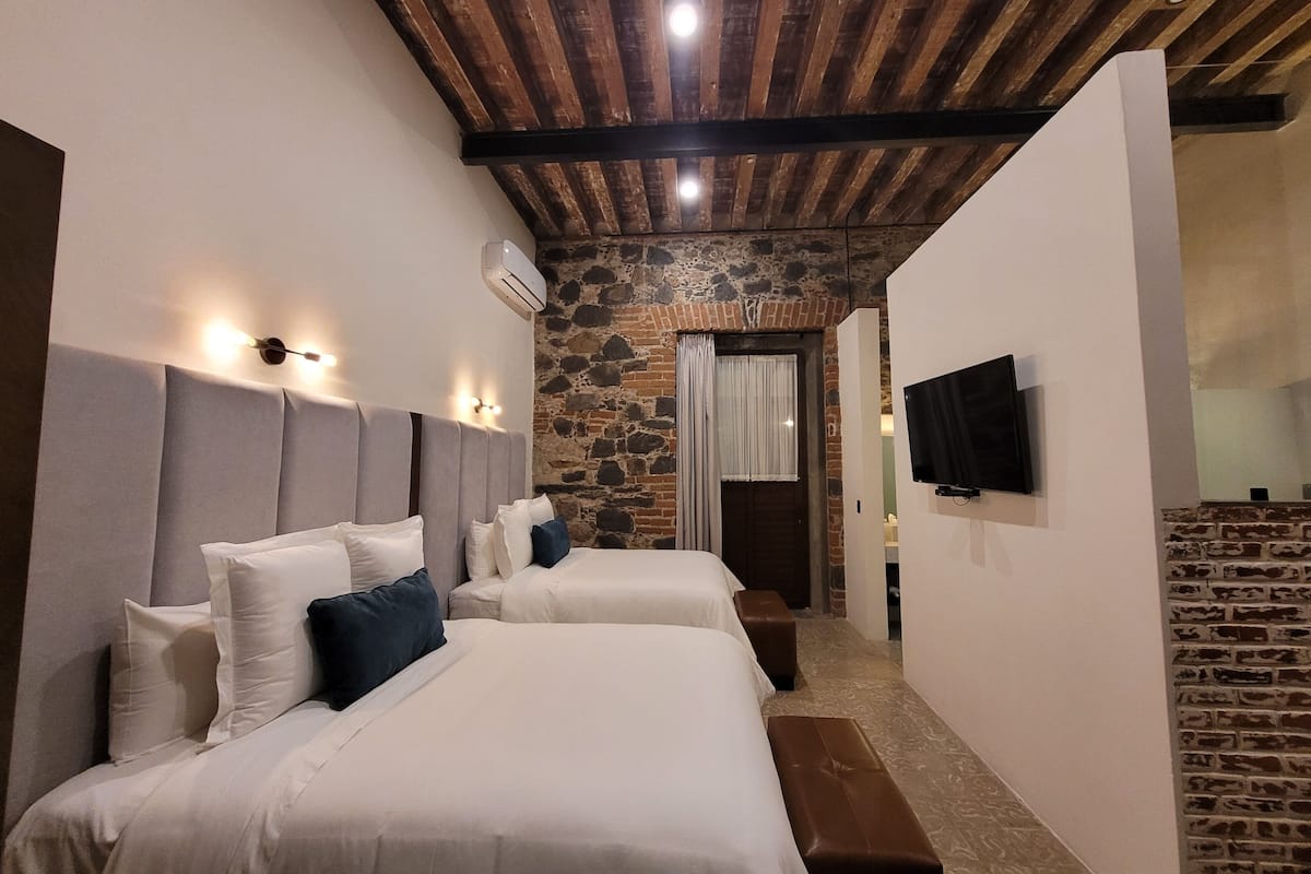 A hotel room with two beds, a stone wall, a wooden ceiling, and a TV mounted on the wall.