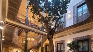 A spacious indoor courtyard with a central tree, stone flooring, and a staircase leading to an upper level. - Milo Collection Hotel (Puebla)