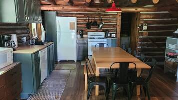 Ein rustikales Blockhaus-Interieur mit einem Holztisch, Stühlen, einer Küchenzeile inklusive Kühlschrank, Herd und hängenden Töpfen.
