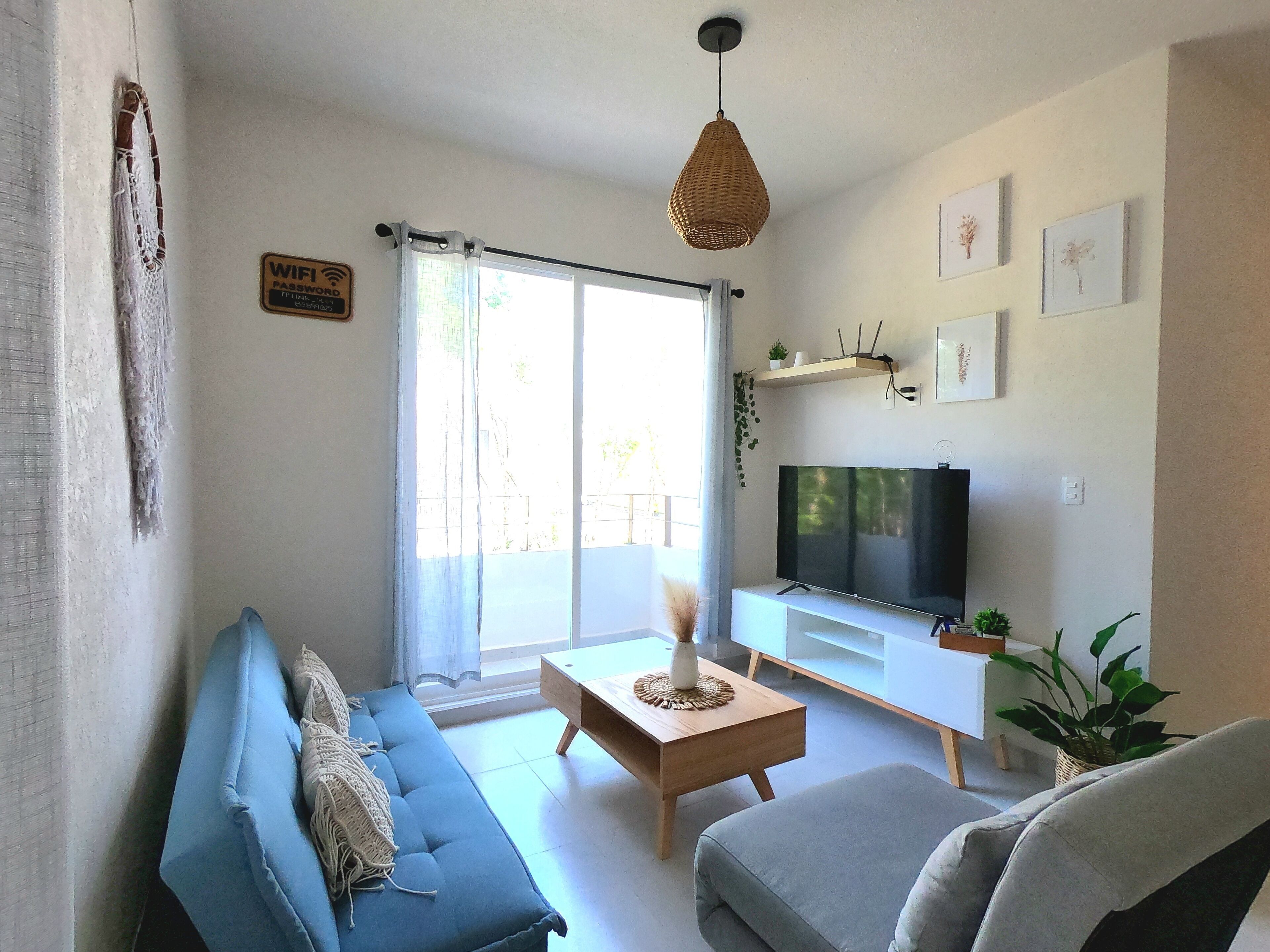 Ett vardagsrum med en blå soffa, en grå fåtölj, ett träbord, en tv och ett stort fönster med gardiner.