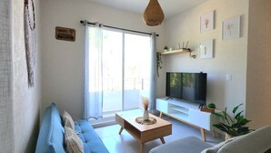 Un salon avec un canapé bleu, un fauteuil gris, une table basse en bois, un téléviseur et une grande fenêtre avec des rideaux.