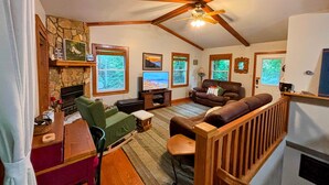 A spacious living room with a stone fireplace, wooden ceiling beams, and large windows.
