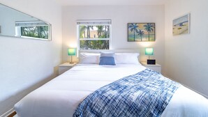 Habitación de hotel ordenada con una cama grande, dos mesitas de noche, un ventanal con vistas a palmeras y dos cuadros colgados en la pared.
