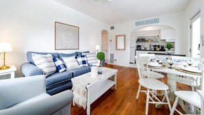 Una sala de estar moderna con un sofá azul, una mesa de centro blanca y un comedor con cuatro sillas blancas y una mesa lista para cuatro comensales.