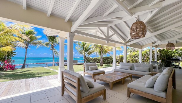A covered outdoor seating area with a view of the ocean, furnished with sofas and tables, and surrounded by palm trees. - Beachfront villa private pool & stunning views (Saint-François)