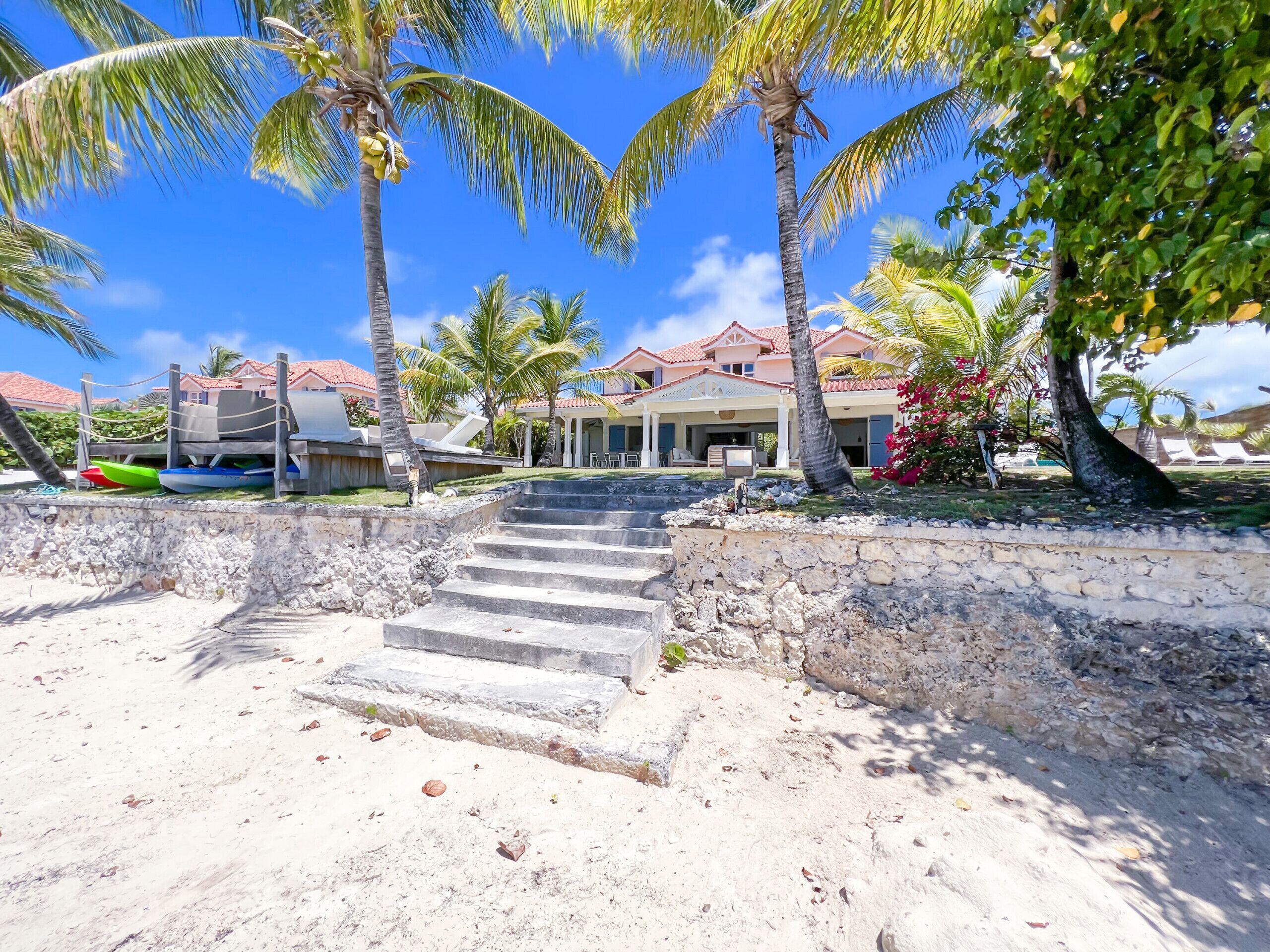 A beachfront property with a stone staircase leading to a covered area, palm trees, and a building with a red-tiled roof.