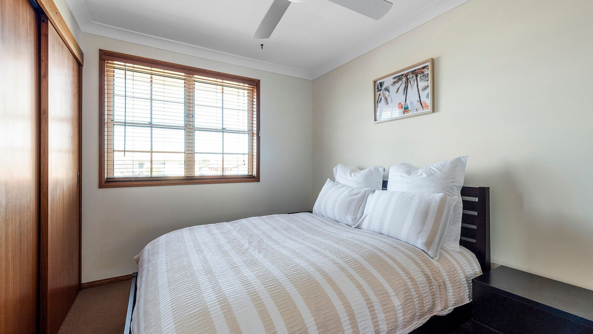 A bedroom with a bed, a window with blinds, a ceiling fan, and a framed picture on the wall.