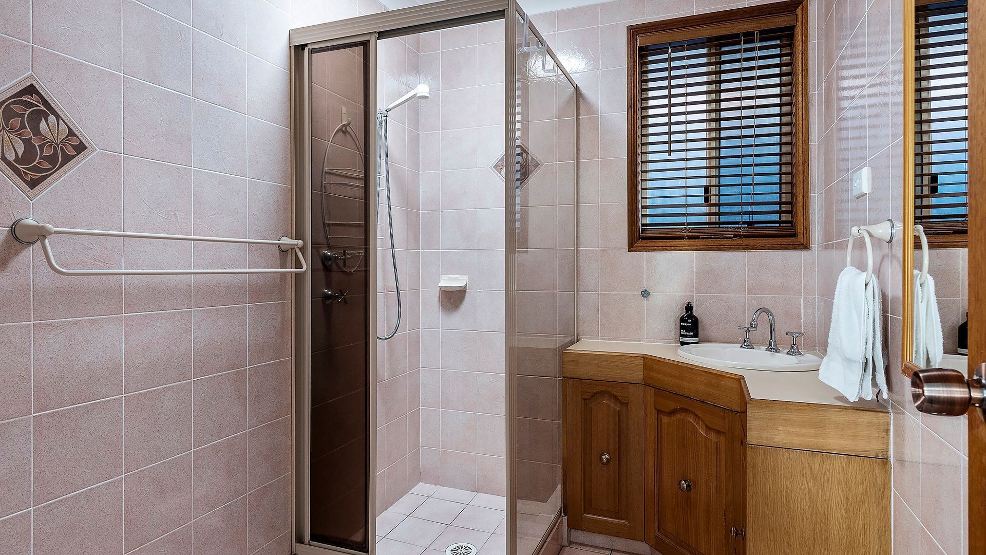 Een badkamer met een douchecabine, een houten wastafelmeubel en een raam met jaloezieën.