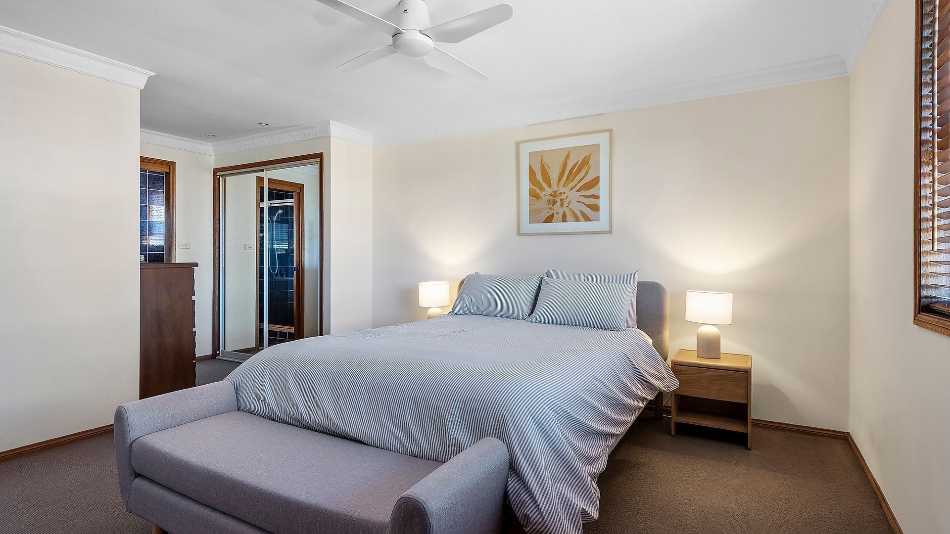 A bedroom with a bed, a chair, a ceiling fan, a nightstand with a lamp, a framed picture on the wall, and a door leading to a bathroom.