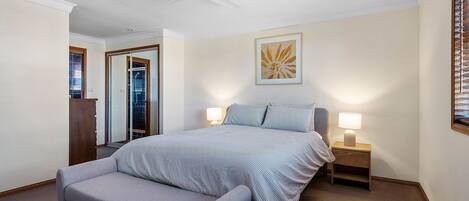 A bedroom with a bed, a chair, a ceiling fan, a nightstand with a lamp, a framed picture on the wall, and a door leading to a bathroom.