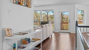 Ein geräumiges Wohnzimmer mit einer Couch, einem weißen Konsolentisch mit Büchern und einem großen Fenster mit Blick auf Bäume und eine Terrasse.