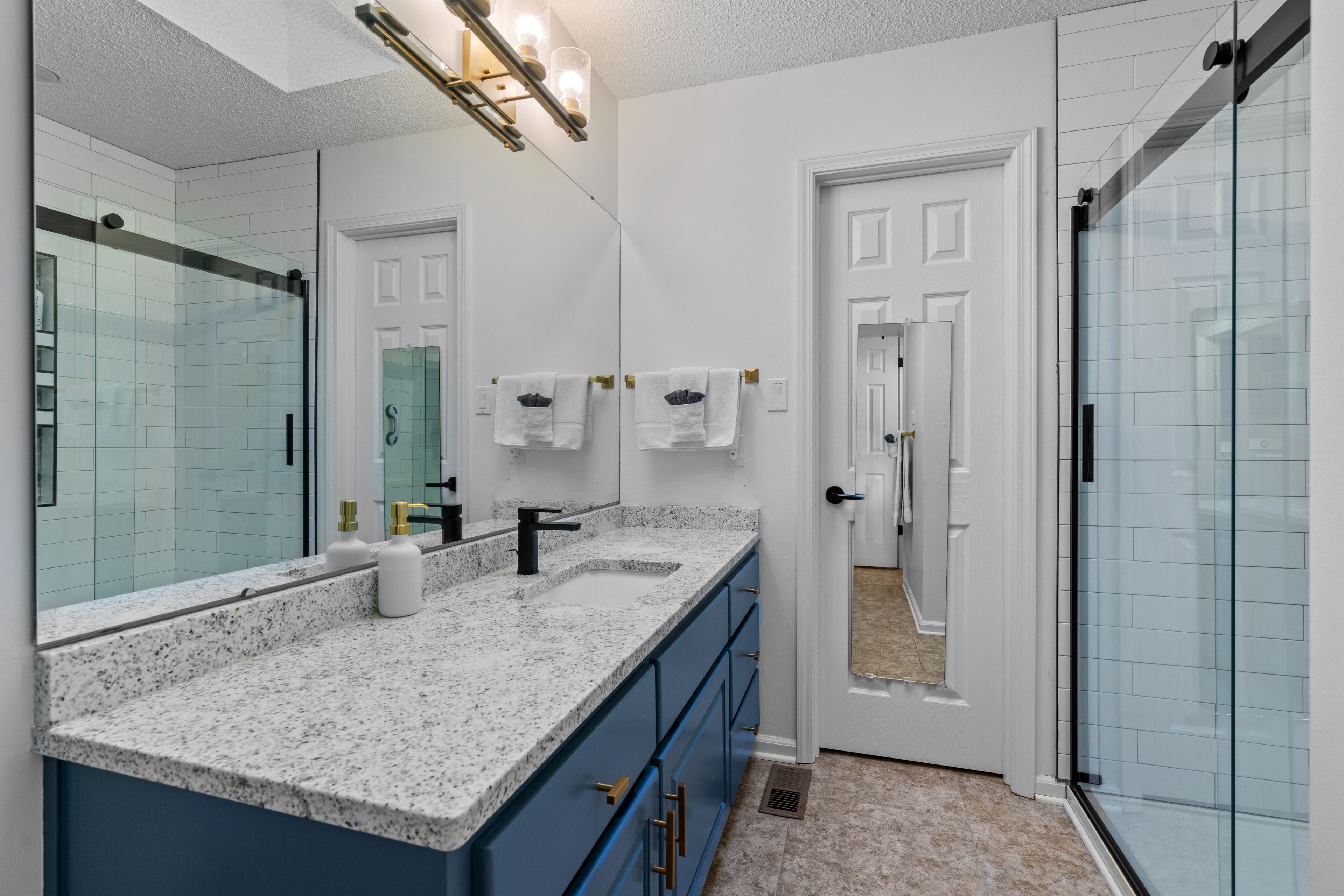 A modern bathroom with a granite countertop, a large mirror, a shower, and a door leading to another room.