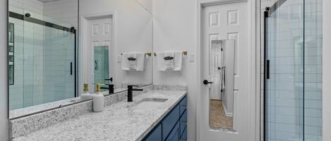 A modern bathroom with a granite countertop, a large mirror, a shower, and a door leading to another room.