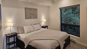 A bedroom with a bed, two bedside tables with lamps, a large window with a view of trees, and a wall-mounted abstract art piece. - J&J`s · Exquisite and brand new 1-bedroom ocean view suite (Bowen Island)