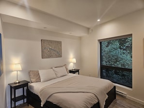 Une chambre Ă coucher avec un lit, deux tables de chevet avec des lampes, une grande fenĂȘtre donnant sur des arbres et une Ćuvre dâart abstraite accrochĂ©e au mur.