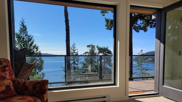 A room with a large window offering a view of the sea and trees. - J&J`s · Exquisite and brand new 1-bedroom ocean view suite (Bowen Island)