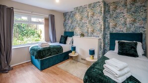 A bedroom with two beds, a window with curtains, a floral wallpaper, and a view of greenery outside.
