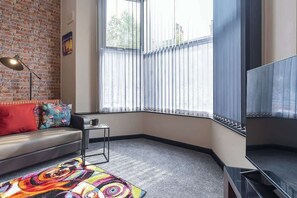 Ein Eckzimmer mit Ziegelwand, einer Couch, einem Fernseher, einem Fenster mit Jalousien und einem farbenfrohen Teppich.