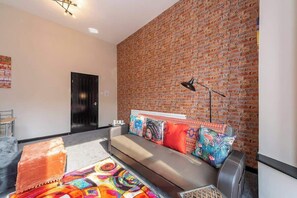 Ein modernes Wohnzimmer mit Backsteinwand, einer Couch mit bunten Kissen, einem gemusterten Teppich und einer schwarzen Stehlampe.