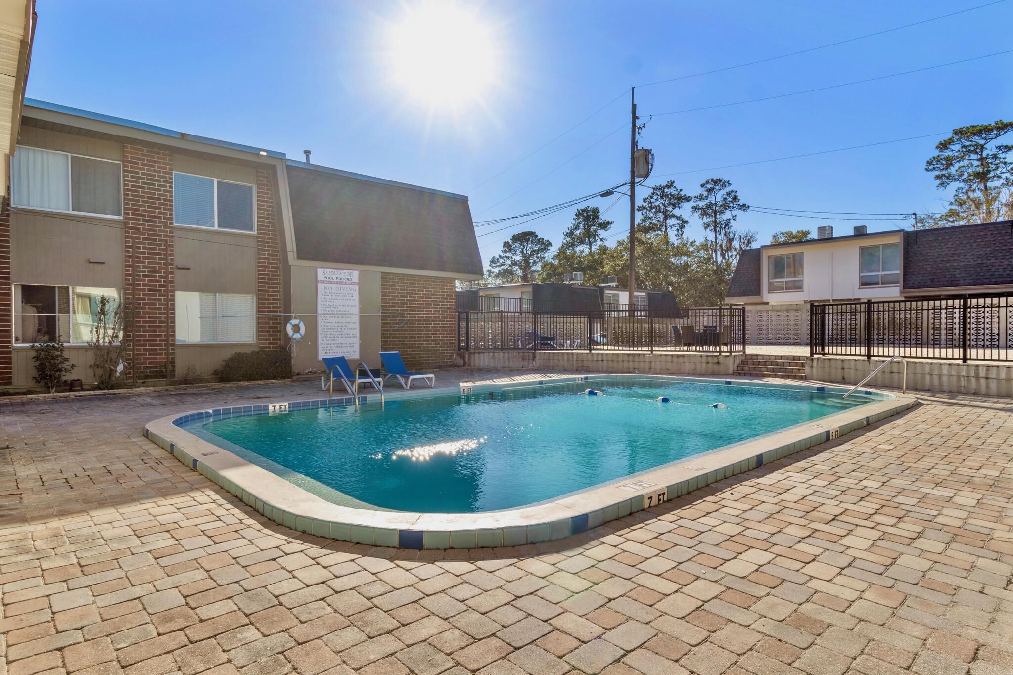 A rectangular swimming pool with a tiled edge, surrounded by a brick patio. There are lounge chairs and a fence enclosing the pool area.