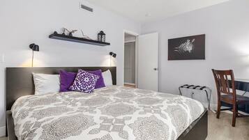 A bedroom with a large bed, two bedside lamps, a chair, a shelf with decorative items, and a zebra artwork on the wall.