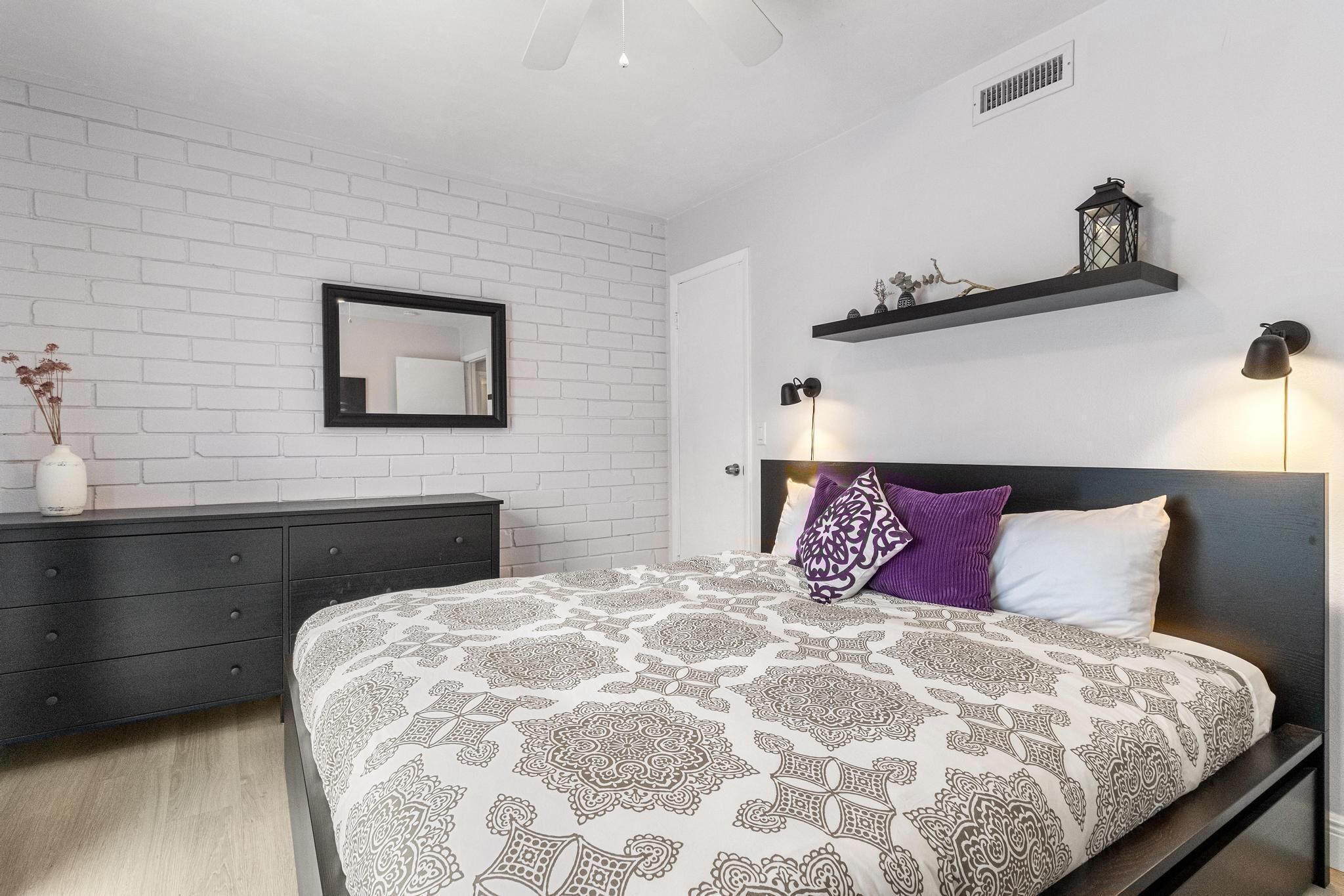 A bedroom with a patterned bedspread, a dark headboard, a white brick wall, a mirror, a dresser, and a shelf with decorative items.