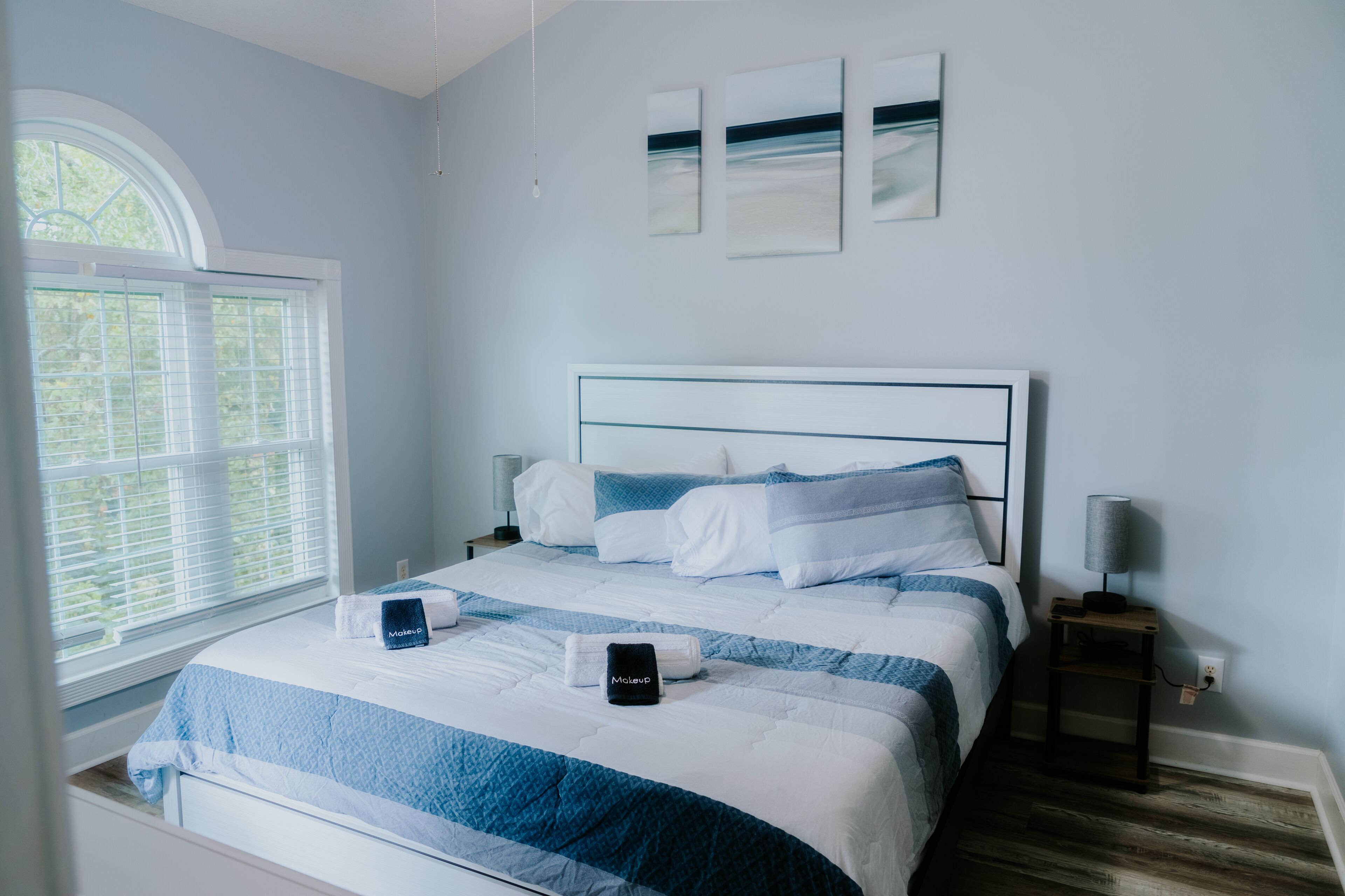 A neatly made bed with a blue and white striped bedspread, a bedside table with a lamp, and a window with blinds.