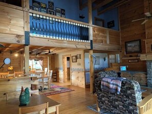 A cozy cabin interior with a wooden floor, a dining area, a couch, and a balcony with a view.