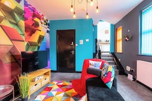 A modern living room with a flat-screen TV, a colorful geometric rug, a red sofa, and a staircase leading to an upper level.