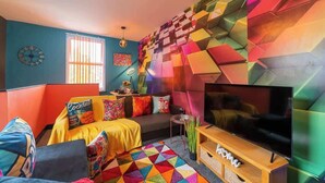 Ein modernes Wohnzimmer mit einem großen Flachbildfernseher, farbenfroher geometrischer Wandkunst und verschiedenen Sitzgelegenheiten, darunter ein Sofa und ein Sessel.