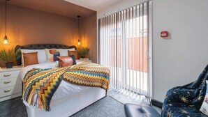 A hotel room with a bed, bedside tables, a chair, a window with blinds, and a red emergency exit sign. - Vibrant Funkily Decorated 2 Bedroom 3 Beds (Kingston upon Hull)
