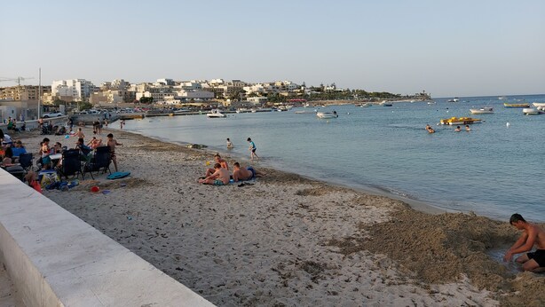 Ein Strand mit badenden Menschen, Booten im Wasser und Gebäuden im Hintergrund.