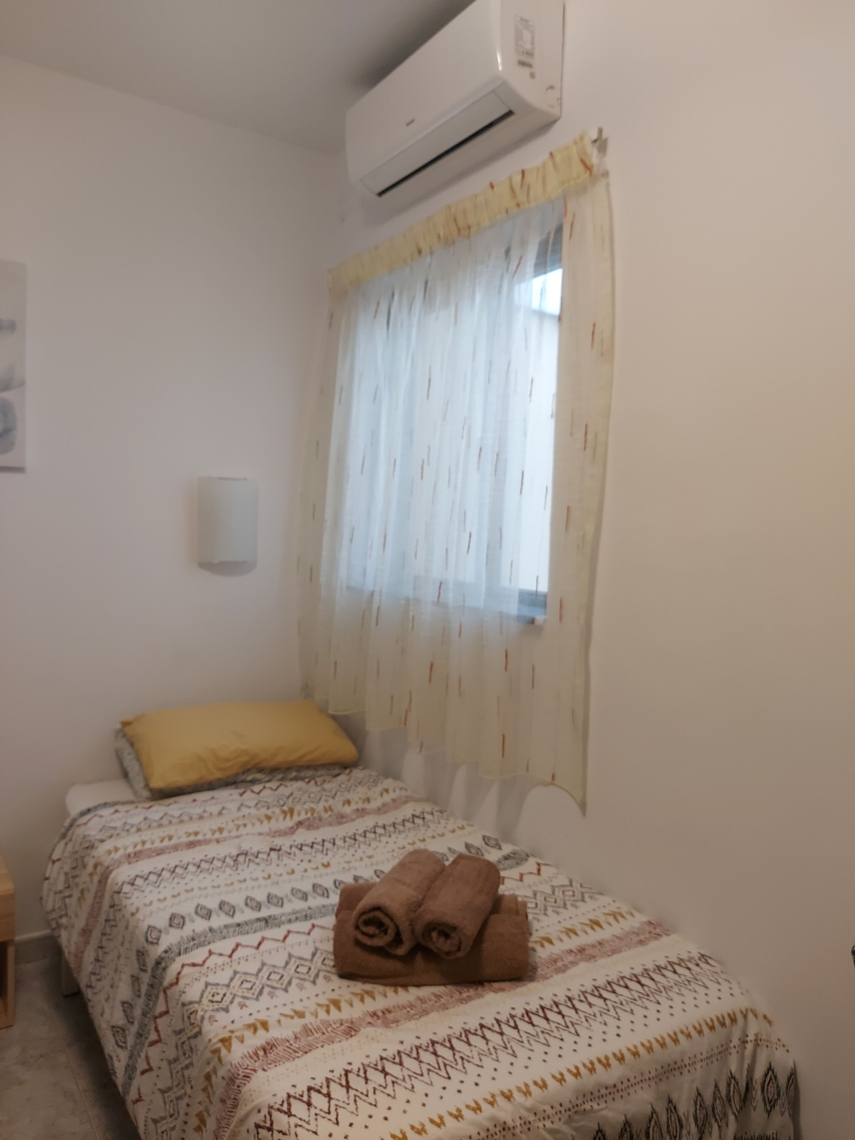 Un dormitorio con cama, ventana con cortinas translúcidas, equipo de aire acondicionado y una lámpara fijada a la pared.