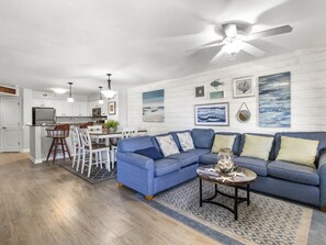 Un salon moderne avec un canapé bleu, un coin repas avec des chaises blanches et une cuisine avec des meubles blancs.