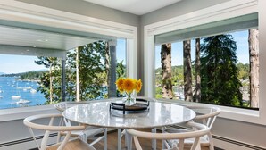 Un espace de restauration moderne, doté d’une table en verre et de chaises blanches, qui offre une vue imprenable sur une magnifique baie, avec des bateaux et de grands arbres.