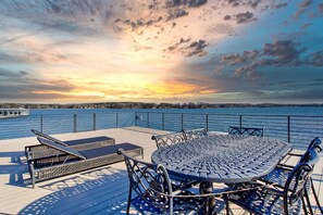 Una terraza con una mesa redonda y sillas, con vista a un cuerpo de agua y un atardecer de fondo.