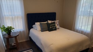 A neatly made bed with a blue headboard, white bedding, and decorative pillows. There is a small wooden table with a plant and a black and white striped pillow. A black and white striped rug is on the floor. - Quiet and Cozy Cottage -10 minutes from the beach! (Corpus Christi)