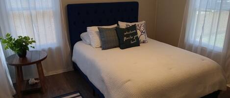 Un letto rifatto con precisione, con una testiera blu, lenzuola bianche e cuscini decorativi. È presente un piccolo tavolino con una pianta e un cuscino a righe bianco e nero. Un tappeto a righe bianche e nere è posto sul pavimento.